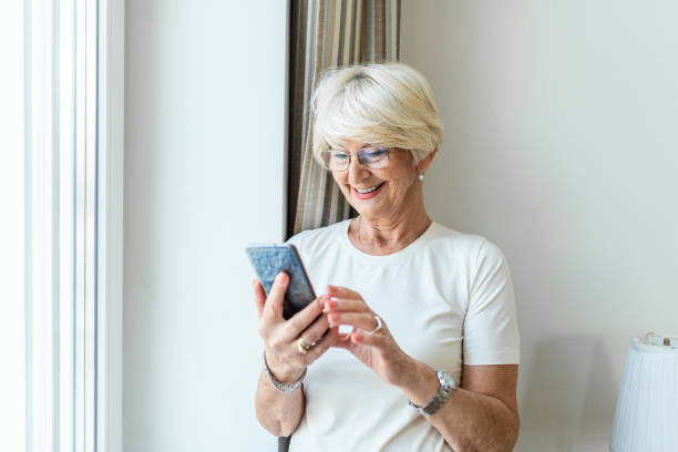 Portrait of cheerful senior woman using smartphone Photo of gray hair woman relaxing at home reading her text messages on her mobile phone with a quiet smile. Senior female texting or playing an online game on smartphone at home. Portrait of cheerful senior woman using smartphone Photo of gray hair woman relaxing at home reading her text messages on her mobile phone with a quiet smile. Senior female texting or playing an online game on smartphone at home. 65 69 years stock pictures, royalty-free photos & images