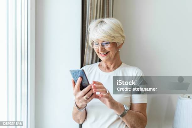 Portrait Of Cheerful Senior Woman Using Smartphone Photo Of Gray Hair Woman Relaxing At Home Reading Her Text Messages On Her Mobile Phone With A Quiet Smile Senior Female Texting Or Playing An Online Game On Smartphone At Home Stock Photo - Download Image Now