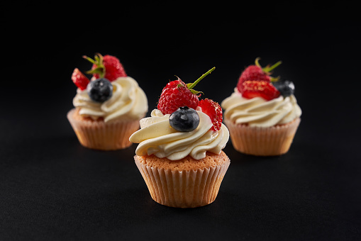 Three fresh tasty cupcakes with white creamy topping in row isolated on black background. Closeup front view of sweet delicious dessert with strawberry, blueberyy and rasberry slices.