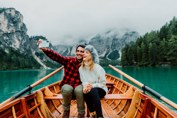 romantic couple of adults in love taking a selfie on a boat visiting an alpine lake at braies italy at autumn fall. couple, wanderlust and travel concept. cold colours. - winter destination imagens e fotografias de stock