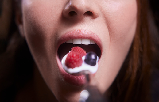 Close-up of young woman mouth eating yogurt with raspberries and blueberries on a silver spoon