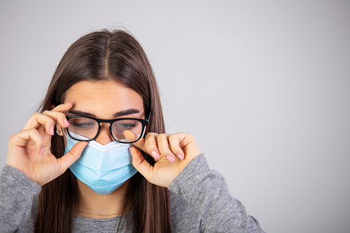 Woman wiping foggy glasses caused by wearing medical mask on gray background. Woman with foggy glasses caused by wearing disposable mask on gray background, space for text