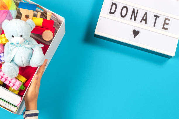 istock Hands holding donation box with toys books clothing and lightbox with text Donate on light blue background. Top view 1289347807