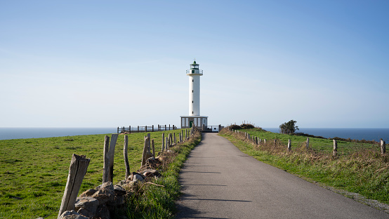 A landscape of a road bordered on the sides of green grass leads to a white lighthouse on the sea coast on a sunny day
