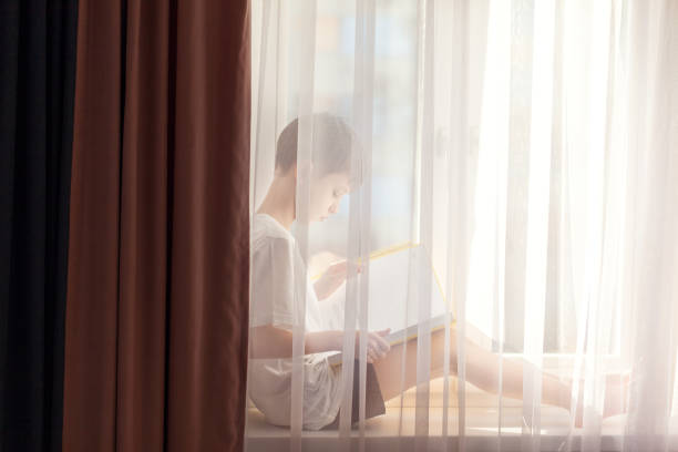 Cute little boy reading book sitting on window at home. Distance learning online education