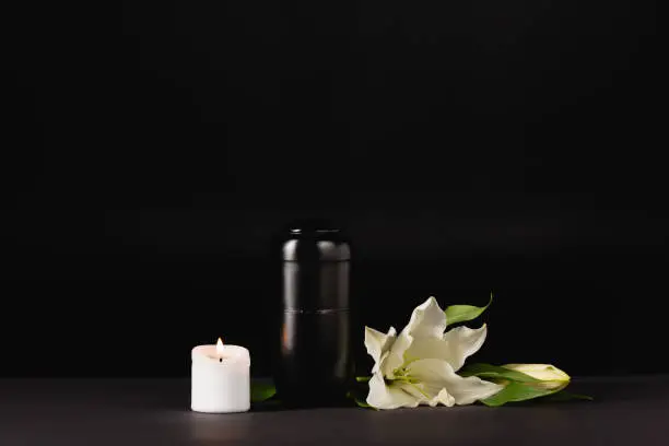 Photo of lily, candle and urn with ashes on black background, funeral concept