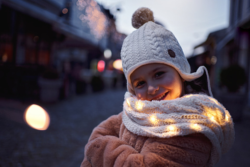 Adorable and excited 5 year old girl wearing scarf with Christmas light around her during dusk at the city