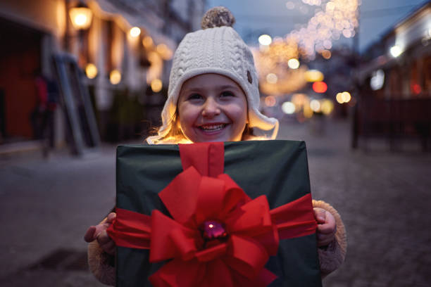 Adorable and excited 5 year old girl holding a Christmas present, wrapped with big red ribbon, and wearing scarf with Christmas light around her during dusk at the city