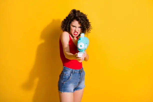 Revenge. Photo of cute aggressive student, girl hold water gun pissed-off aiming shot young brother wear jeans shorts red singlet unclothed shoulders isolated bright yellow color background