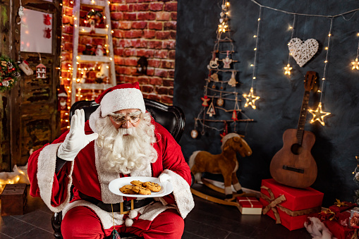 Cheerful Santa Claus holding plate with cookie while sitting at his chair.