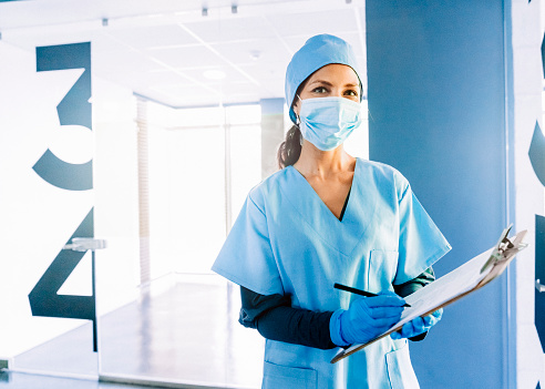 Portrait of female nurse holding clipboard with medical record. Healthcare worker is in blue scrubs at hospital. She is wearing face mask and glove during pandemic.