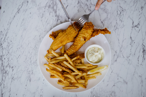 An Asian Chinese woman eating fish and chips meal using kitchen knife and fork in a cafe