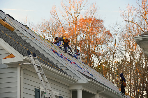 Ocean Pines, MD - Dec 3, 2020:  A roofing crew three adult men of Hispanic ethnicity from central America replacing the shingles on a residential building, one of the most dangerous jobs in America,  as the sun sets on a winter day in early