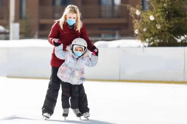 young mother teaching her little daughter ice skating at outdoor skating rink. Family wearing a medical mask during COVID-19 coronavirus enjoy winter on ice-rink outdoors
