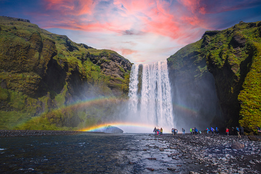 Group of tourists looking at the beautiful rainbow at the impressive Skogarfoss waterfall in southern Iceland, photo taken in summer on a beautiful sunset