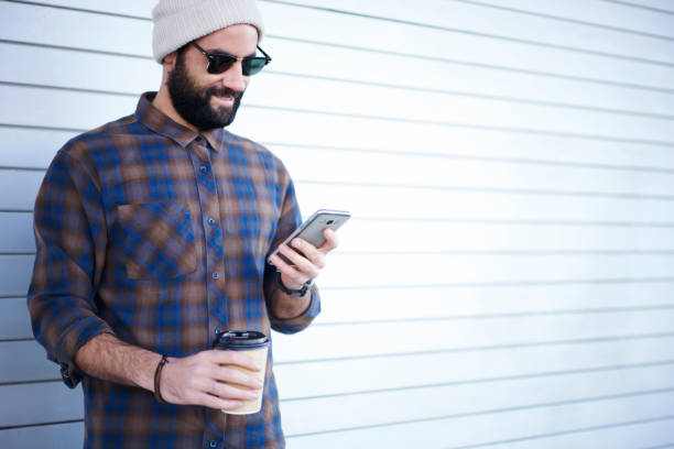 istock Crop young bearded man with cup of coffee using smartphone 1289393882