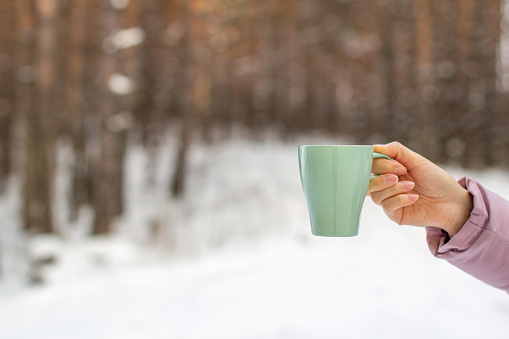 A young woman holds a glass of hot coffee or tea in the forest in winter. Snowy winter and hot drink to keep warm. Cup of hot coffee or tea