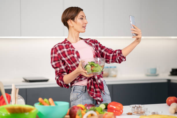 istock Pretty housewife taking selfie or making a video call using her smartphone while cooking fresh salad wearing a plaid shirt with a bob hair style. Healthy food leaving - vegan concept 1289399567
