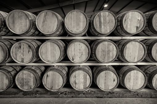 A black and white image of a front on view of a row of stacks of traditional full whisky barrels, set down to mature, in a large warehouse facility