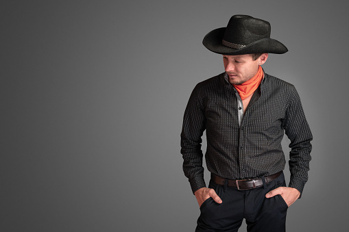 Cowboy in a black shirt with his hands in his pants pockets. Unshaven smiling man in a hat. Macho guy isolated on gray background. Looks down and to the side. Western American culture concept