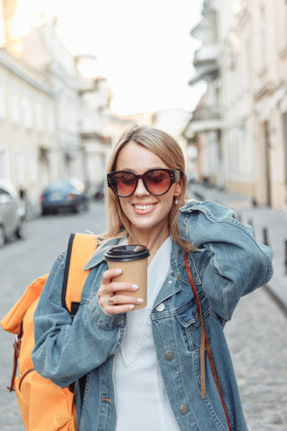 istock Nice beautiful millennial tourist with backpack in jeans jacket holds coffee cup and discover European city at holiday travel. 1289405828