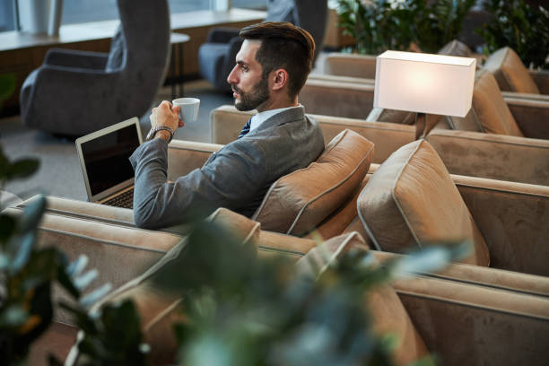 Serious handsome person drinking coffee in the business lounge Attractive man in a suit holding a cup and looking away while being alone with his laptop gate stock pictures, royalty-free photos & images