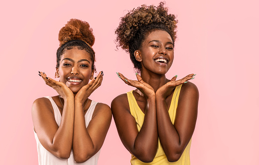Happy overjoyed two afro women smile broadly, looking at camera, posing on pink pastel studio background.