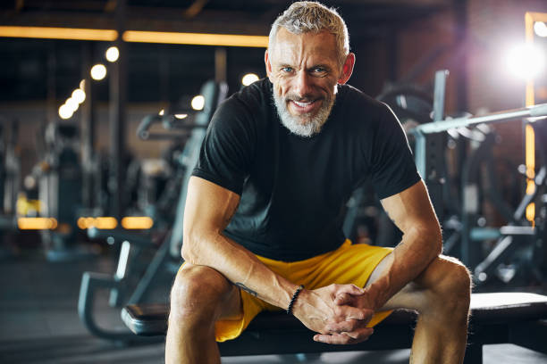 Sportsman with a happy smile sitting on the weight bench Front view of a smiling pleased bearded gray-haired male athlete with interlocked fingers looking ahead mature men stock pictures, royalty-free photos & images