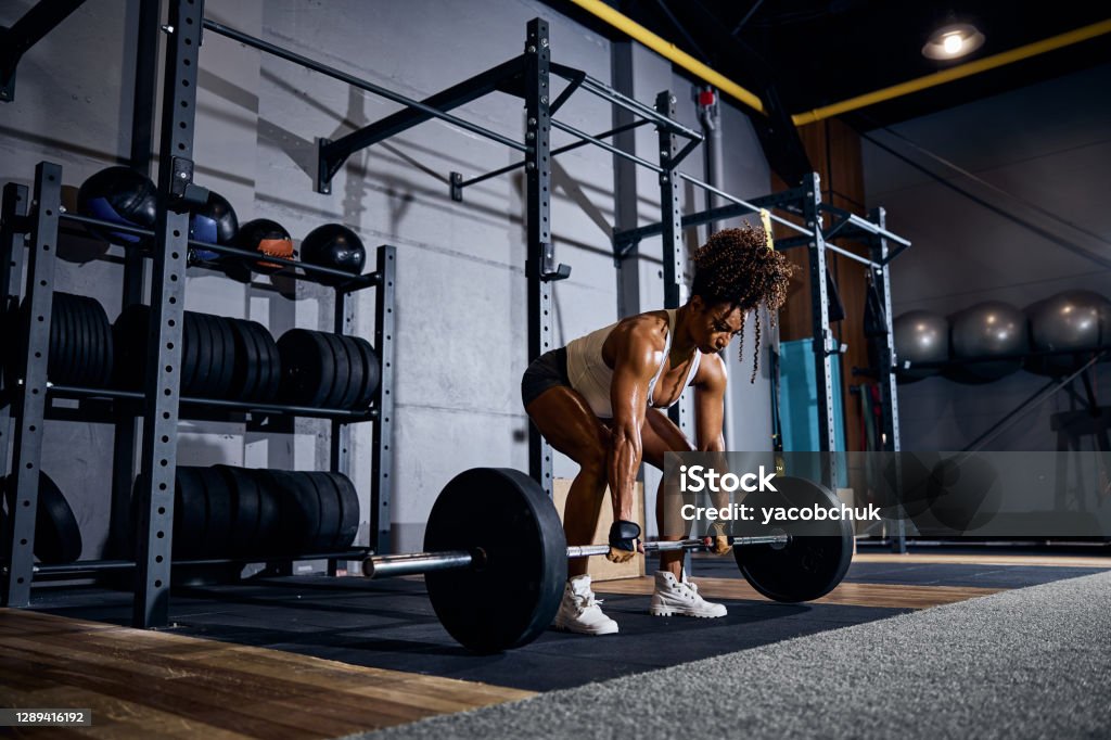 Strong young woman performing the deadlift exercise Serious muscular African American lady lifting the loaded barbell off the ground with both hands Weightlifting Stock Photo