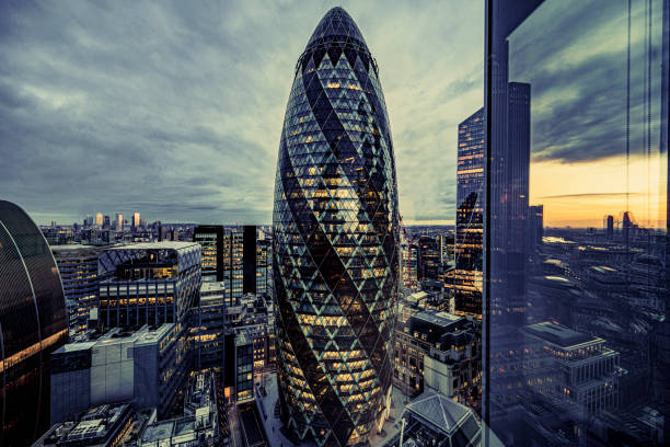 Illuminated office buildings in City of London at sunset Elevated perspective of landmark modern architecture with warm light shining in windows and sunset reflecting in foreground glass beneath cirrus clouds. london gherkin at night stock pictures, royalty-free photos & images