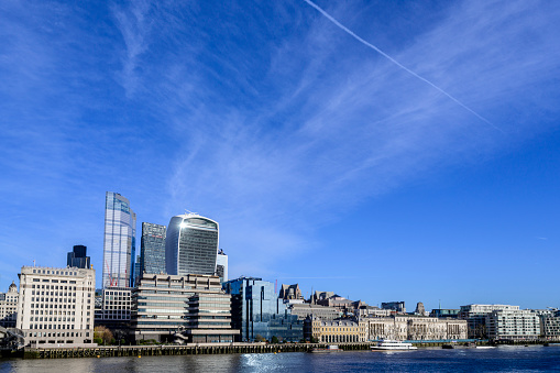 Wide angle view from opposite shore of City of London skyline with landmark buildings and cirrus clouds and vapor trail overhead.