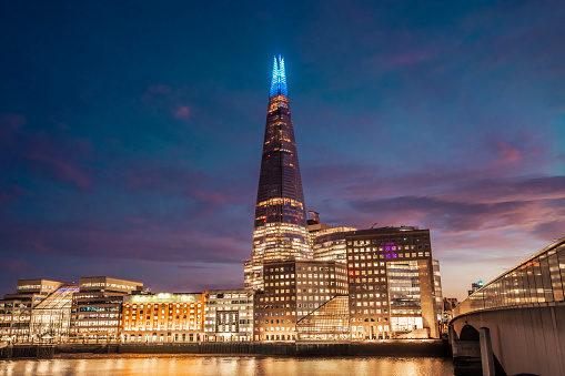 View across Thames River of pyramidal tower in neo-futuristic style with spire illuminated in blue to honor nation’s healthcare workers in time of COVID-19.