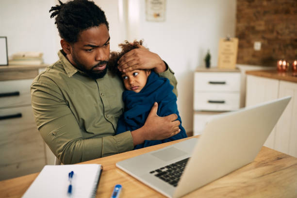 istock Black single father using laptop while holding his ill small daughter at home. 1289419165