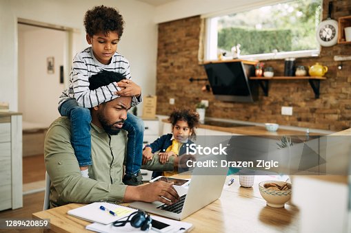 istock Black stay at home father working on laptop while his kids are demanding his attention. 1289420859