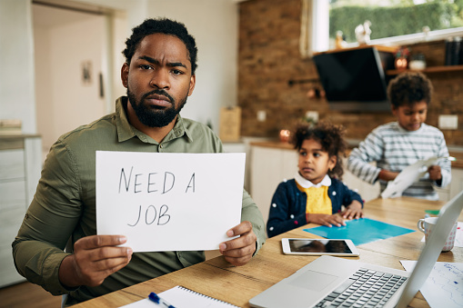 Black stay at home father holding placard with 'need a job' inscription while babysitting his children at home.