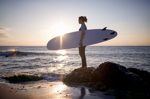 silhouette of female surfer standing on the beach at sunrise, she holding surfboard and looking away.