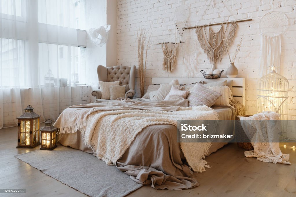 Comfortable apartment with bright and cozy interior design Side view of lovely bedroom with plaid and pillows on comfortable bed, home decor and cushions on soft armchair in white interior design in bohemian style Bedroom Stock Photo