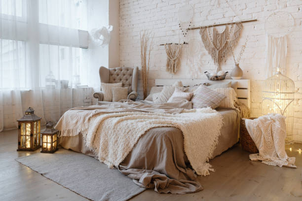 Comfortable apartment with bright and cozy interior design Side view of lovely bedroom with plaid and pillows on comfortable bed, home decor and cushions on soft armchair in white interior design in bohemian style macrame photos stock pictures, royalty-free photos & images