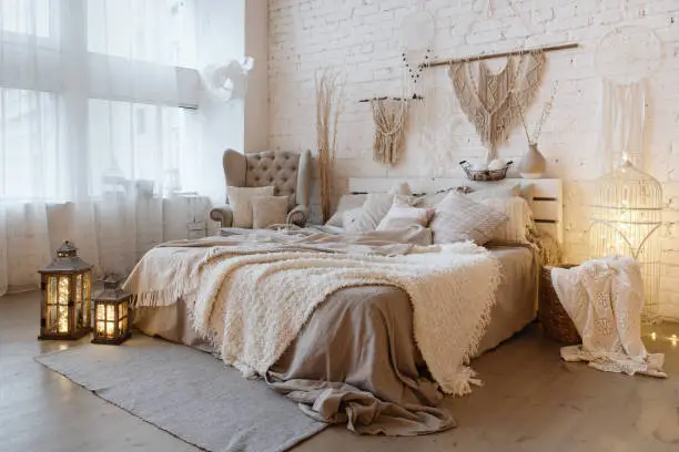 Side view of lovely bedroom with plaid and pillows on comfortable bed, home decor and cushions on soft armchair in white interior design in bohemian style