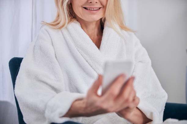 istock Happy smiling woman in white plush bathrobe using cell phone and internet in spa center 1289422528