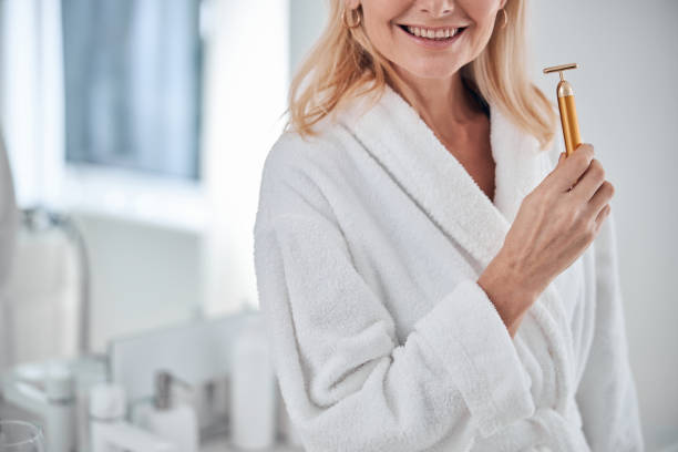 hermosa mujer encantadora en bata de felpa blanca usando rodillo facial mientras está de pie en la habitación en el interior - quartz caucasian one person energy fotografías e imágenes de stock