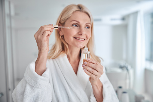 Close up portrait of charming woman holding bottle with dropper near face while applying cosmetic facial oil on face with pipette in bathroom