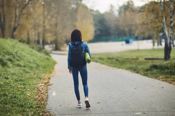 A brunette girl with a bag on her back goes to the Park to make sports. She is holding a yoga Mat. The view from the back. The fall of the year.