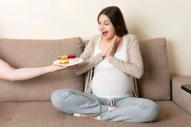 Photo of Husband offers his pregnant wife tasty cakes relaxing on the sofa at home. Future father takes care about expecting mother