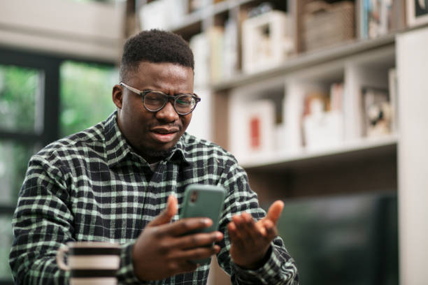 Young African American man having reading bad news on his smart phone Annoyed young African American man sitting at home, reading some bad news on his smart phone using a mobile app and expressing his displeasure guy looking at cell phone stock pictures, royalty-free photos & images