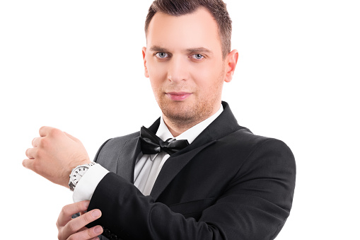 Portrait of a confident attractive young man in black suit with bow tie, adjusting the cufflinks on his white shirt, looking at the camera, isolated on a white background.