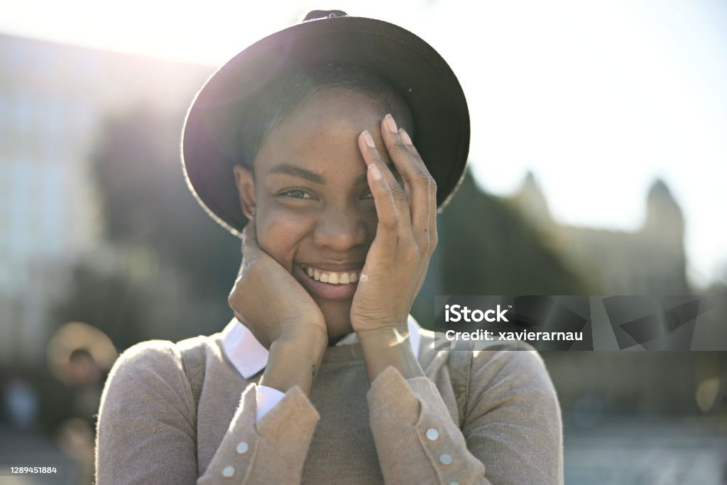 Portrait of Shy Black Woman Guarding the Joy on Her Face Partial front view of mid 30s woman standing outdoors on sunny day in hat and casual clothing cradling her face as she smiles at camera with great happiness. Shy Stock Photo