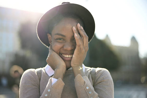 Portrait of Shy Black Woman Guarding the Joy on Her Face Partial front view of mid 30s woman standing outdoors on sunny day in hat and casual clothing cradling her face as she smiles at camera with great happiness. shy stock pictures, royalty-free photos & images