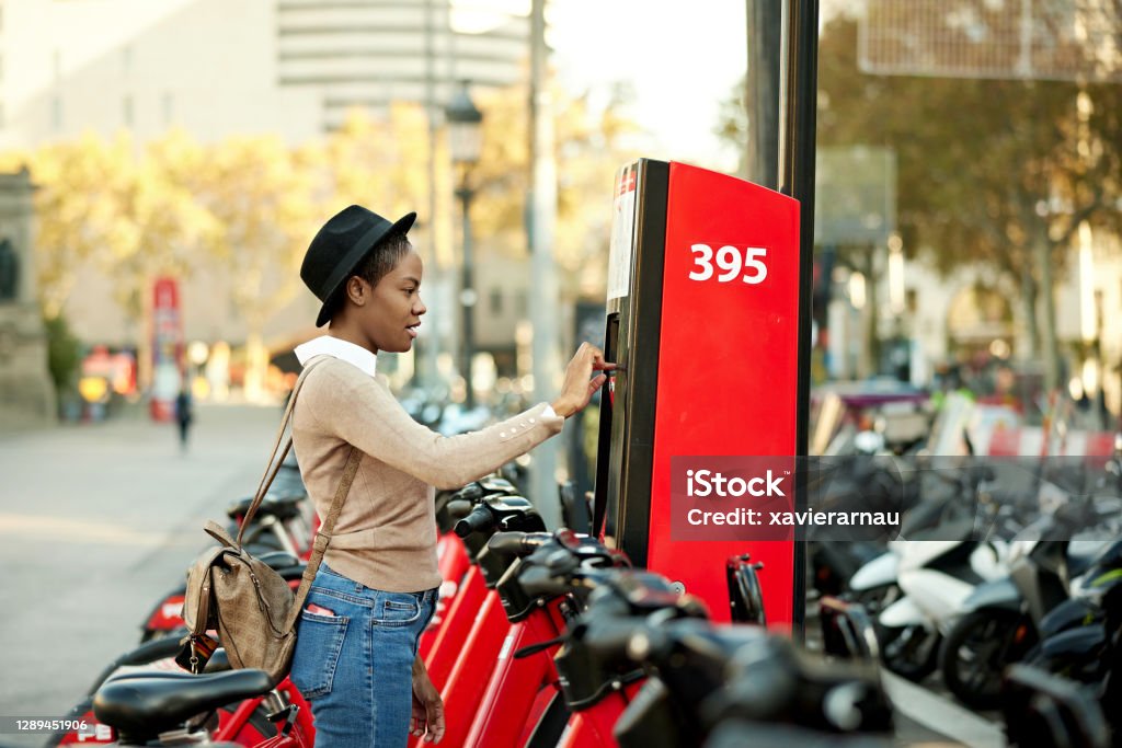 Socially Responsible Mid 30s Black Woman Renting Bicycle Partial side view of woman in hat and casual clothing standing at self-service bicycle sharing kiosk paying for her rental in urban setting. Bicycle Stock Photo