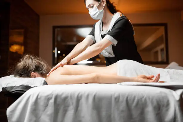 Professional female masseur in facial mask doing massage to a client at Spa salon. Business during the epidemic concept